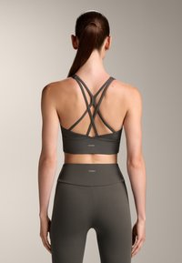 Temno siva športna bralette s križnimi naramnicami na hrbtu, v kombinaciji z visokimi pajkicami. Ima gladko teksturo in znamčni detajl.