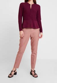 Frau trägt eine bordeauxrote, strukturierte, zum Peplum geschnittene Reißverschlussjacke, staubrosa maßgeschneiderte Hose und schwarze, Riemchen-Absatzsandalen, stehend vor weißem Hintergrund.