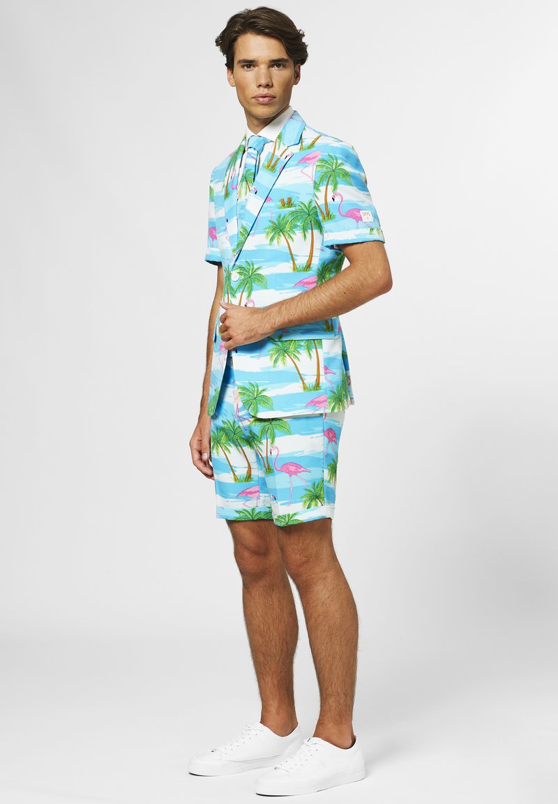 Costume bleu clair à manches courtes et shorts, orné d'un motif floral tropical avec des palmiers et des flamants roses. Paire de baskets blanches.
