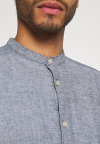 Light blue linen shirt with a stand-up collar, featuring small round buttons and a textured fabric detail near the button placket.