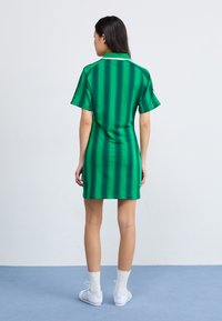 Rochie verde cu dungi, cu mâneci scurte și guler; are un design strâns. Poartă șosete albe și adidași. Vedere din spate.