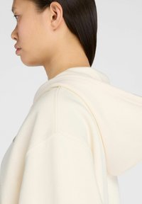 Sweatshirt à capuche crème avec une coupe décontractée, présentant un petit logo sur le devant et un tissu doux et texturé.