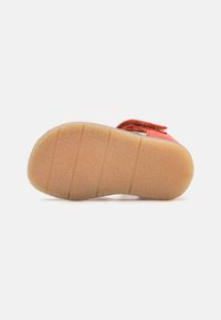 Röd mockaskor med en texturerad beige gummisula, som har horisontella spår för grepp och en mjuk yta. Vinkelsnabb för stängning.