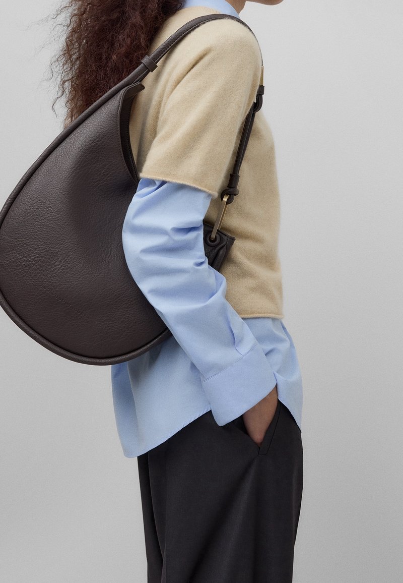 Femme portant un pull beige à manches courtes par-dessus une chemise bleue, pantalon noir, portant un grand sac à bandoulière en cuir marron foncé sur fond gris.