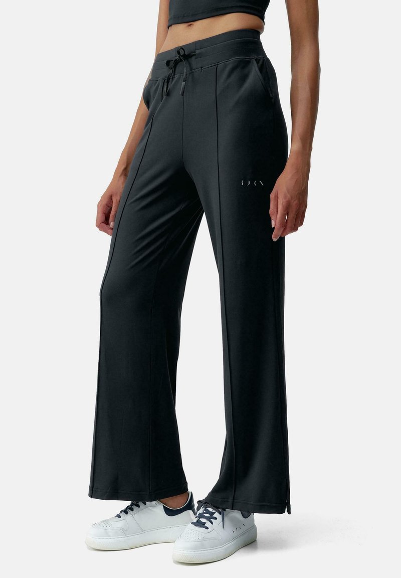 Pantaloni atletici neri a gamba larga con una trama liscia, vita elastica, coulisse e sottili dettagli laterali. Abbinati a sneakers bianche.