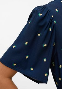 Blouse bleu marine avec manches courtes bouffantes, présentant un motif répétitif de citrons jaune et vert sur un tissu doux. Fronces délicates aux épaules.