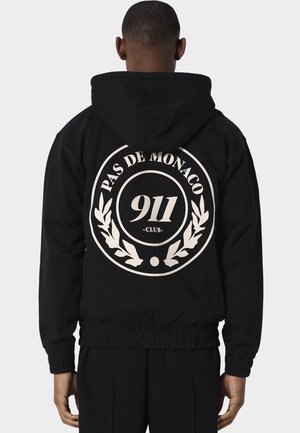 Persoon draagt een zwart jack met capuchon met een groot wit cirkelvormig "PAS DE MONACO 911 CLUB" logo op de achterkant, staand tegen een effen achtergrond.