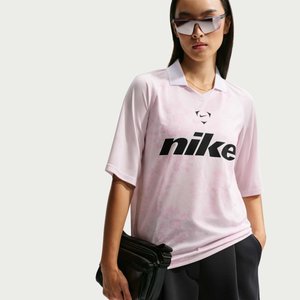 Ung kvinde iført oversized lyserød Nike-shirt, store sportslige solbriller, holder sort clutch med hånden i lommen på neutral baggrund.