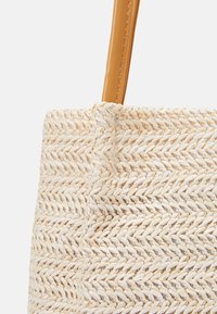 Zbliżenie beżowej torby z wikliny z wzorem w zygzak i jasnobrązowym skórzanym paskiem na białym tle.