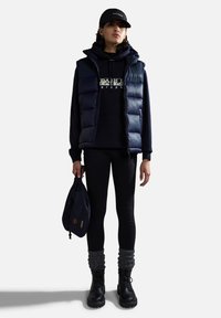Marineblauwe puffer vest over een marineblauw hoodie, zwarte leggings en zwarte veterschoenen. Accessoires omvatten een donkere pet en een marineblauwe trekkoordtas.