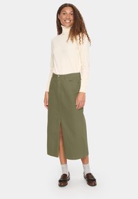 Olivno zelena, dolg kroj krilo, izdelano iz trdnega blaga, z odprtino spredaj, v kombinaciji s kremasto turtlenek majico in rjavimi mokasini.