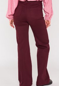 Pantaloni svasati bordeaux in tessuto liscio con tasche posteriori. Cuciture visibili e dettaglio della cintura piatta. Modello che indossa una maglietta a righe.