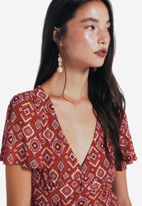 Rotes Kleid mit Muster, tiefem V-Ausschnitt, kurzen Ärmeln und plissierter Textur. Es weist geometrische Designs in Weiß, Gelb und schwarzen Akzenten auf.