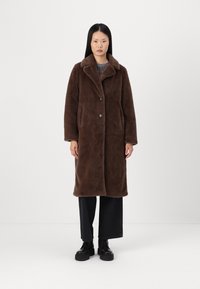 Cappotto in pile marrone con colletto, due bottoni e tasche laterali. Tessuto liscio, maniche lunghe, lunghezza fino al ginocchio, abbinato a pantaloni neri a gamba larga.