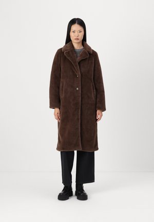 Cappotto in pile marrone con colletto, due bottoni e tasche laterali. Tessuto liscio, maniche lunghe, lunghezza fino al ginocchio, abbinato a pantaloni neri a gamba larga.