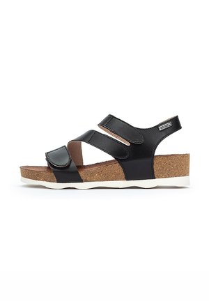 Sandalias sintéticas negras con dos correas ajustables de Velcro, una plantilla de corcho y una suela de goma blanca. Diseño plano y abierto para mayor comodidad.