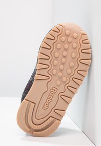 Podeszwa zewnętrzna buta Reebok, wykonana z beżowej gumy z okrągłymi i prążkowanymi wzorami. Ciemny materiał wierzchni widoczny wzdłuż krawędzi.