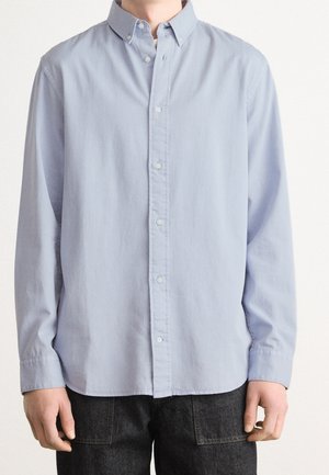 Homme portant une chemise bleu clair à manches longues boutonnée et un pantalon foncé, debout devant un fond blanc uni.