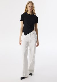 Schwarzes, tailliertes T-Shirt mit leicht asymmetrischem Schnitt, kombiniert mit weißen, weit geschnittenen Jeans mit fransigen Kanten und zwei vorderen Taschen.