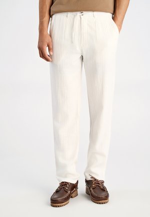 Witte, geplooide broek met een tailleband met trekkoord, gemaakt van lichtgewicht stof. Draagt zich met bruine leren schoenen met veters en textuur op de zolen.
