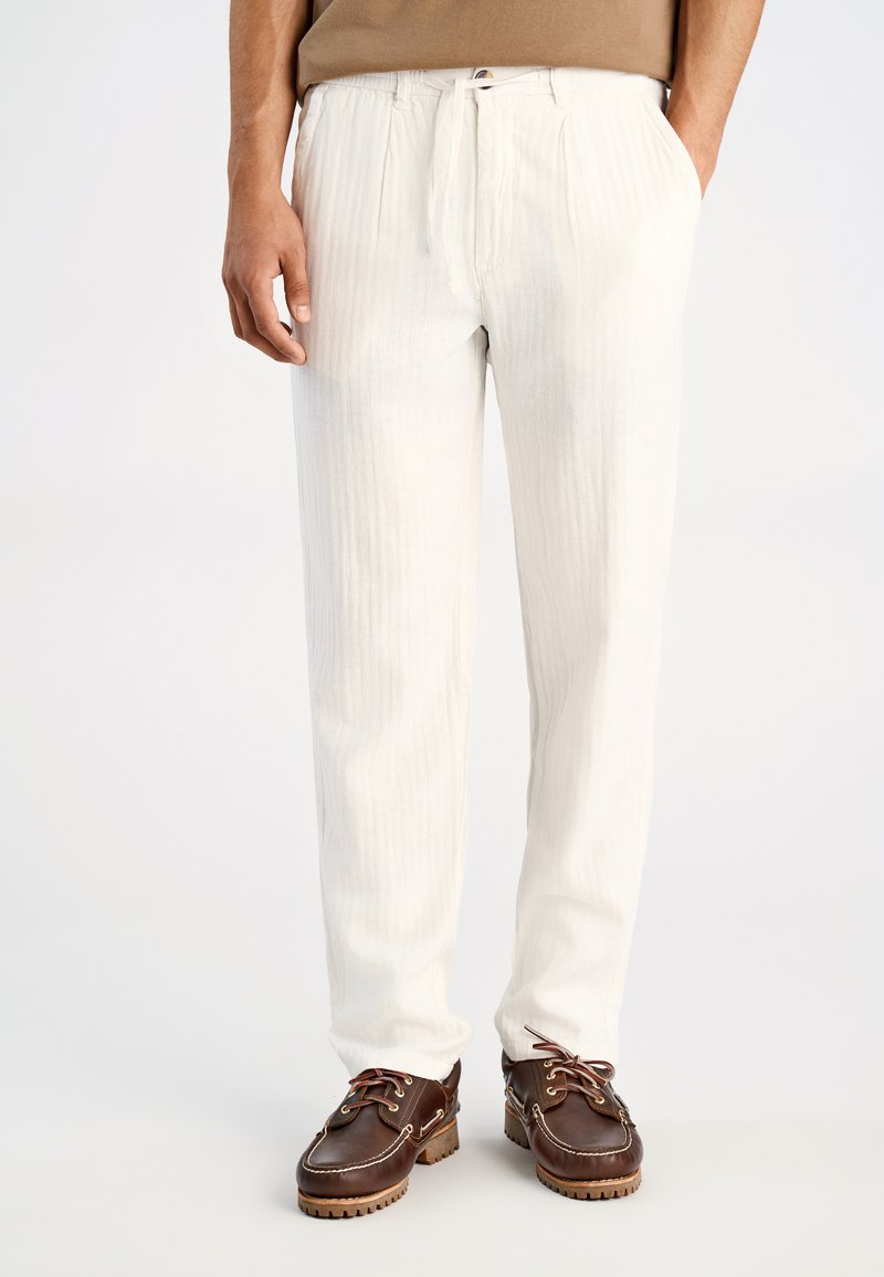 Witte, geplooide broek met een tailleband met trekkoord, gemaakt van lichtgewicht stof. Draagt zich met bruine leren schoenen met veters en textuur op de zolen.