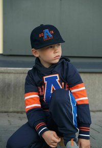 Laivaston sininen huppari, jossa oransseja ja valkoisia raitoja, sinisellä kirjaimella "A" ja numerolla "2016". Musta lippalakki kirjailtuine yksityiskohtineen. Harmaat tennarit.