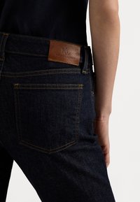Temno modri jeans iz denima z teksturiranim zaključkom, s kontrastnim rumenim šivanjem in rjavim usnjenim oznako, na kateri je vtisnjen "Ralph Lauren."