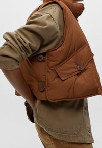 Brown puffer vest with a side pocket, large buttons, and a padded texture, layered over a beige long-sleeve shirt.