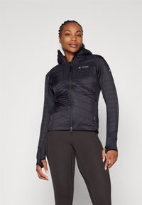 Giacca nera isolante con cappuccio, caratterizzata da chiusura con zip, motivo a zigzag, maniche aderenti e foro per il pollice, abbinata a leggings scuri.