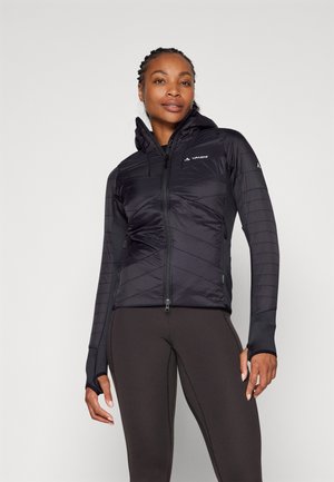 Chaqueta negra aislante con capucha, que presenta un cierre de cremallera, patrón en forma de chevrón, mangas ajustadas y orificios para los pulgares, combinada con leggings oscuros.