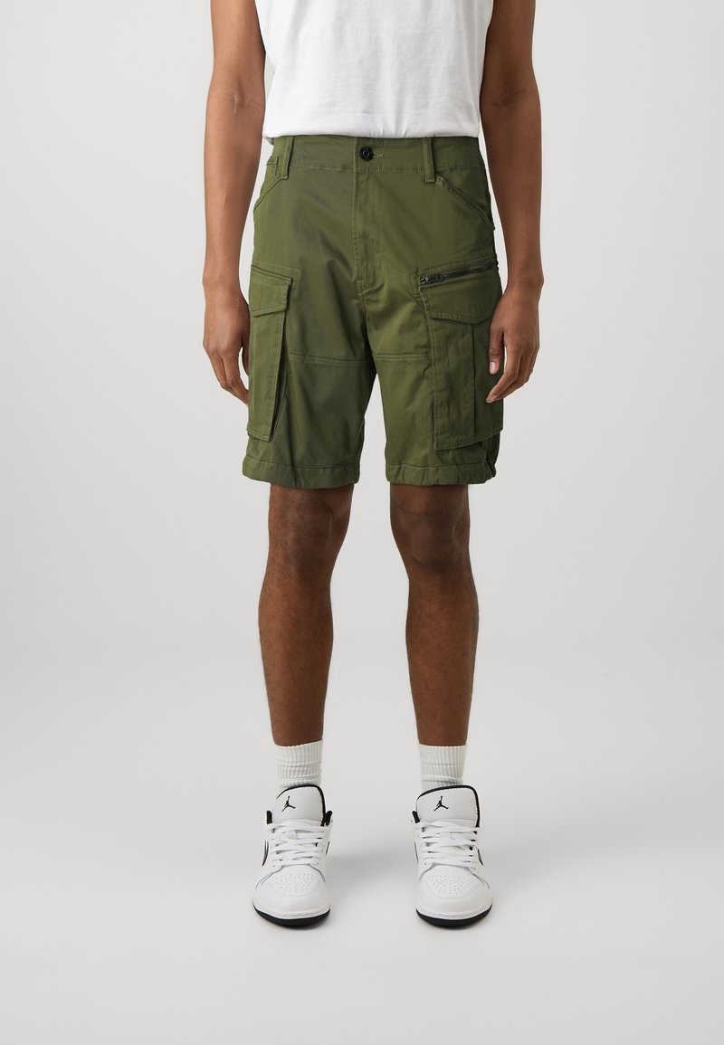 Man i olivgröna cargo-shorts, vita strumpor och vita hög-top sneakers, stående mot en enfärgad ljusgrå bakgrund.