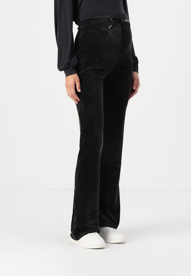 Pantalons en velours noir avec une forme évasée, dotés d'une taille à cordon de serrage et d'une texture lisse. Portés avec des baskets blanches pour le contraste.