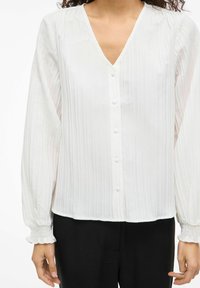 Hvid bluse med lodrette striber, V-udskæring, fem knapper foran og rynkede manchetter. Letvægts, tekstureret stof, afslappet pasform.
