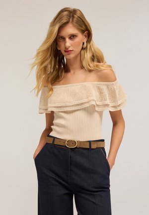 Jonge vrouw met lang blond haar, gekleed in een beige off-shoulder top met ruffles, gouden oorbellen, een gouden riem en donkerblauwe broek, met haar handen in de zakken.