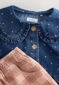 Jeanskleid mit kragenförmigem Ausschnitt, blauer Stoff mit rosa Punkten, versehen mit braunen blumenförmigen Knöpfen, kombiniert mit hellkarierten Hosen.
