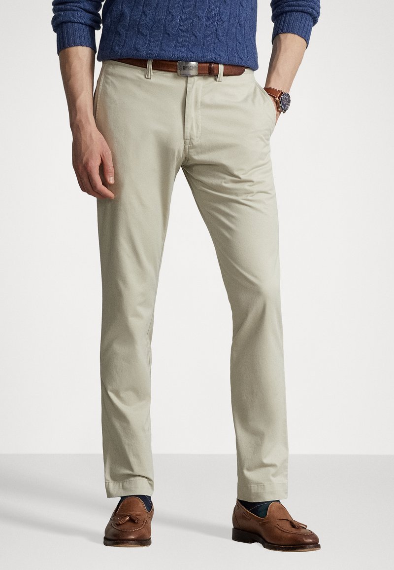 Beige bomullskinoer med rette ben, sidelommer og et brunt lærbelte. Kombinert med brune loafers og en blå kabelstrikket genser.