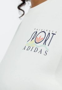 Bijela pamučna majica s višebojnim logotipom: "Originals Sport" s naglaskom u tri pruge i stiliziranim dizajnom planinske valovite linije.