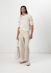 Pull en maille avec un motif ajouré en crème, pantalon beige et baskets blanches avec des semelles brunes, tenant un sac fourre-tout beige.