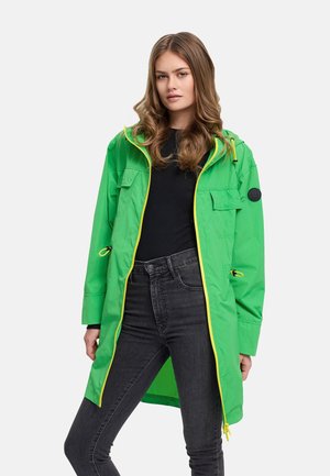 Jonge vrouw die een felgroene gewatteerde jas met gele ritssluitingdetails draagt, een zwart shirt en donkergrijze spijkerbroek, staand tegen een witte achtergrond.