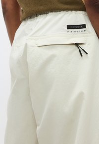 Ανοιχτό μπεζ σορτς με ελαστική μέση, με φερμουάρ πίσω τσέπη και διακριτική υφή. Το λογότυπο στην τσέπη γράφει «IT'S OUT THERE.»