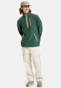Pullover in pile verde con colletto alto, dettagli beige e chiusura a bottoni a pressione. Abbinato a pantaloni cargo beige e sneakers.