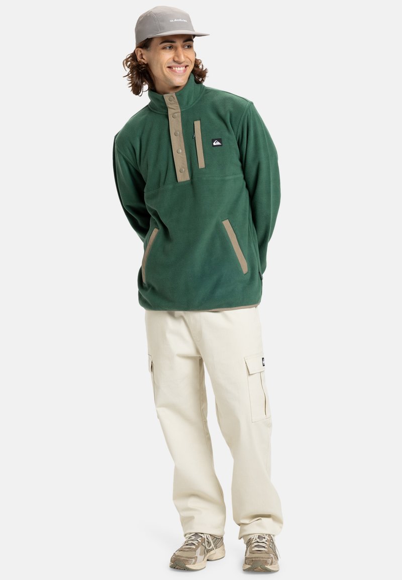Pullover in pile verde con colletto alto, dettagli beige e chiusura a bottoni a pressione. Abbinato a pantaloni cargo beige e sneakers.