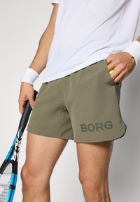 Žalios sportinės šortai su šoninėmis kišenėmis ir subtiliu tamsiai žaliu „BORG“ logotipu. Derinama su balta marškinėliais ir laikoma teniso raketė.