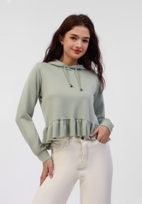 Mintgrønn hettegenser med volangkant, lange ermer og snøring. Kombineres med lyse jeans, har en lomme. Glatt tekstur.