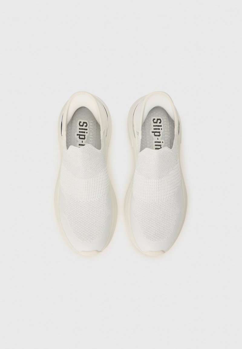 Para białych sneakersów typu slip-on z teksturowanym cholewką i wyściełanymi podeszwami, widziana z góry na białym tle.