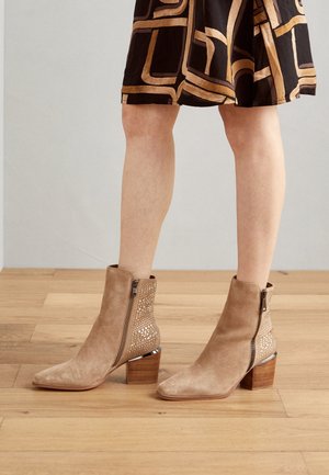 Ben iført beige ruskindsstøvler med blokhæl og lynlåse, kombineret med et sort og beige geometrisk mønstret nederdel.