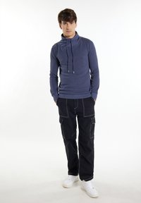 Marineblauer Hoodie mit hohem Kragen und Kordelzügen, aus strukturiertem Stoff, kombiniert mit schwarzen Cargo-Hosen, die weiße Nähte und Taschen haben. Weiße Sneakers.