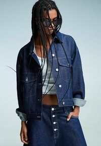 Jeans jakna z temnim pranjem, skrajšano zasnovo, srebrnimi detajli in kontrastnim šivanjem, v kombinaciji z lahko majico brez rokavov in usklajenim krilom.