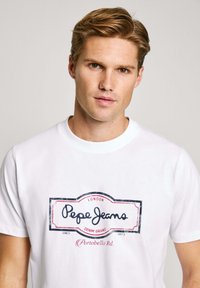 White cotton t-shirt featuring a graphic logo in blue and red, stating "Pepe Jeans London Denim Brand" with a vintage-style border.
