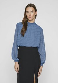 Niebieska zwiewna bluzka z wzorem w zygzak, z wysokim dekoltem i bufkami, zestawiona z czarną spódnicą z bocznym rozcięciem.